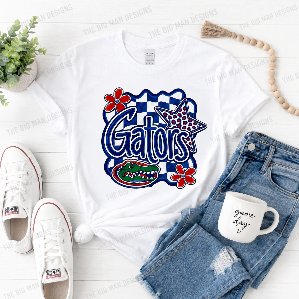 A white graphic T-shirt laid flat on a white wood background, styled with blue denim shorts, white sneakers, a white mug, a small plant, and a wooden bead garland. The shirt features a large Gators graphic with red flowers, a blue-and-white checkerboard outline, a star, and an alligator.