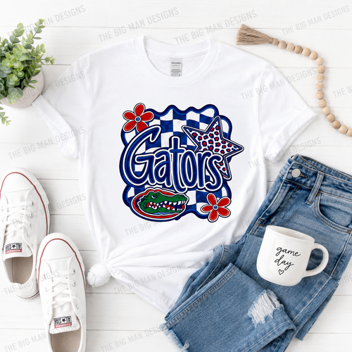 A white graphic T-shirt laid flat on a white wood background, styled with blue denim shorts, white sneakers, a white mug, a small plant, and a wooden bead garland. The shirt features a large Gators graphic with red flowers, a blue-and-white checkerboard outline, a star, and an alligator.