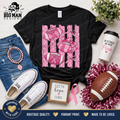 A black graphic T-shirt laid flat on a light wood background with styled accessories around it, including blue jeans, white sneakers, a pink tumbler, a football, tinsel, a small framed sign reading 'faith hope love,' and a pink ribbon. The shirt graphic shows pink footballs and pink polka-dot stripes.
