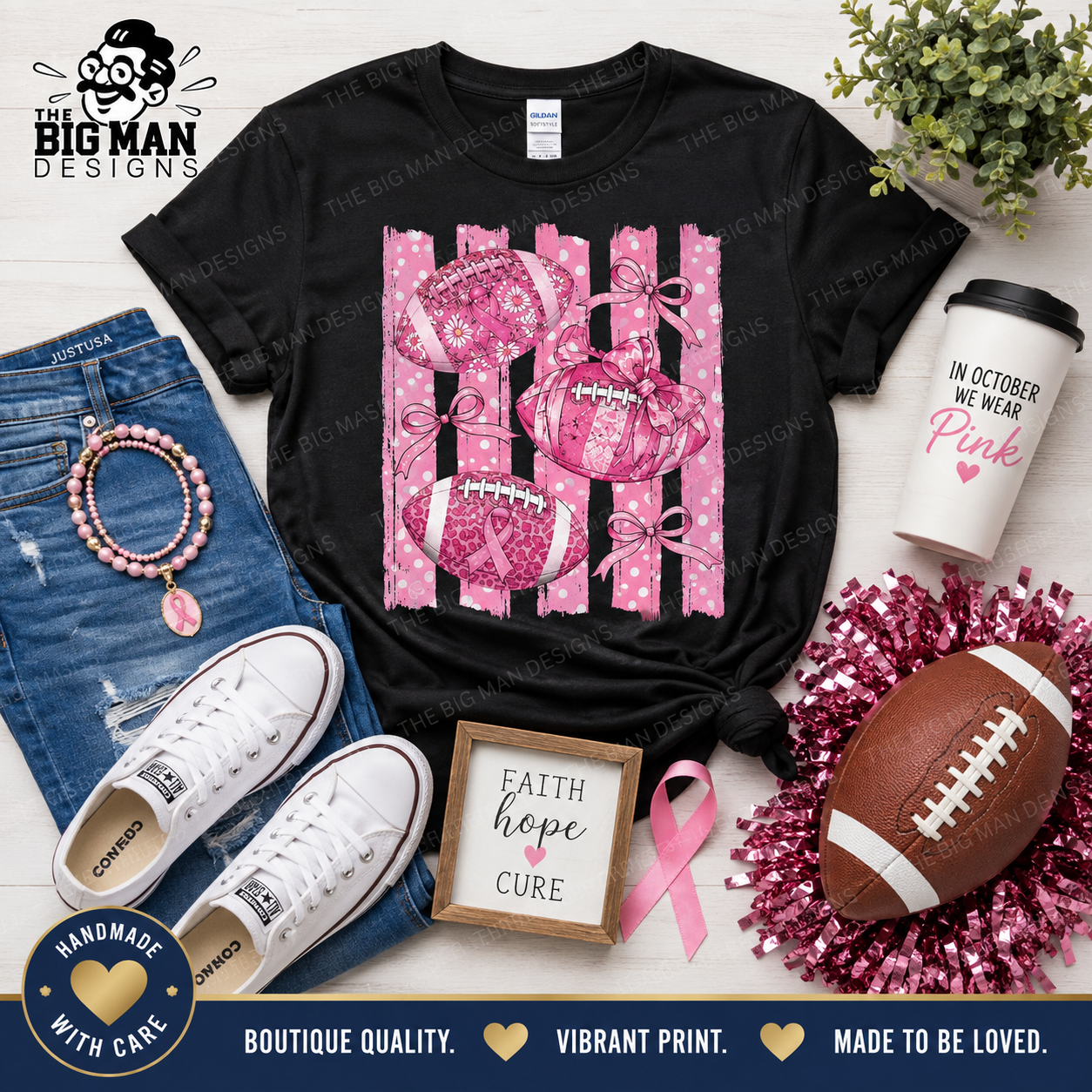 A black graphic T-shirt laid flat on a light wood background with styled accessories around it, including blue jeans, white sneakers, a pink tumbler, a football, tinsel, a small framed sign reading 'faith hope love,' and a pink ribbon. The shirt graphic shows pink footballs and pink polka-dot stripes.