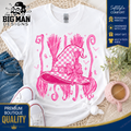 A white graphic t-shirt featuring a pink witch hat design with a checkered pattern, bow, and broomsticks. The t-shirt is styled in a flat-lay arrangement with blue jeans, white sneakers, a pink mug, and a wicker mat, with promotional badges from 'The Big Man Designs' visible.