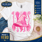 A white graphic T-shirt laid flat on a woven rug with jeans, sneakers, a pink mug, and small greenery around it. The shirt features a large pink witch hat illustration with broomsticks and a bow.