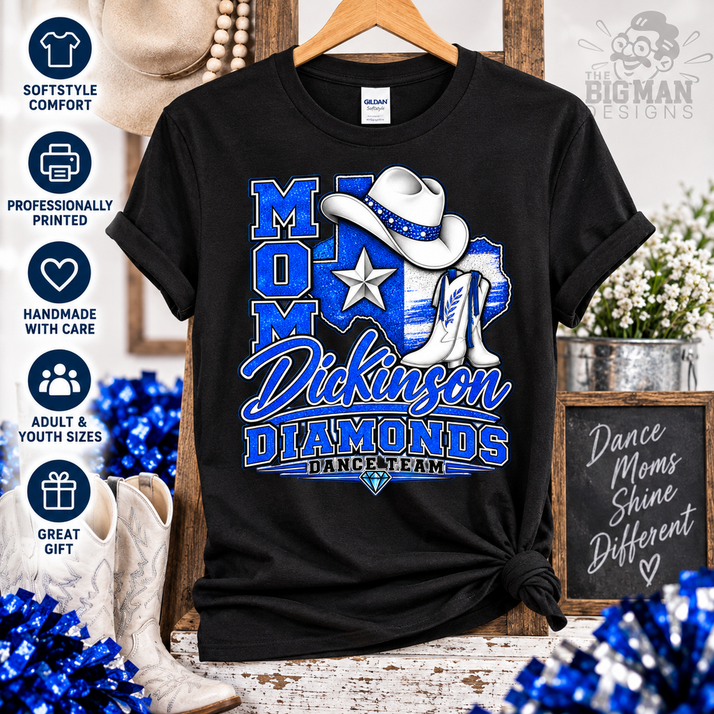 A black short-sleeve graphic T-shirt hanging on a wooden hanger, featuring a western-themed design with the text 'MOM Dickinson DIAMONDS DANCE TEAM' in blue and white, a cowboy hat, boots, and a star. The shirt is shown in a styled rustic setting with decorative props, including blue pom-poms, cowboy boots, flowers, and a sign.