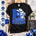 A black short-sleeve graphic T-shirt hanging on a wooden hanger, featuring a western-themed design with the text 'MOM Dickinson DIAMONDS DANCE TEAM' in blue and white, a cowboy hat, boots, and a star. The shirt is shown in a styled rustic setting with decorative props, including blue pom-poms, cowboy boots, flowers, and a sign.