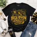 A black Gildan t-shirt with a gold and yellow graphic of a gator and the text 'DICKINSON Gators GOLD OUT', styled in a flat lay with white sneakers, blue jeans, a black tumbler, a gold chain, and a plant on a white wood background.