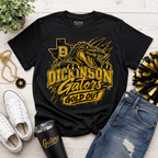 A black Gildan t-shirt with a gold and yellow graphic of a gator and the text 'DICKINSON Gators GOLD OUT', styled in a flat lay with white sneakers, blue jeans, a black tumbler, a gold chain, and a plant on a white wood background.