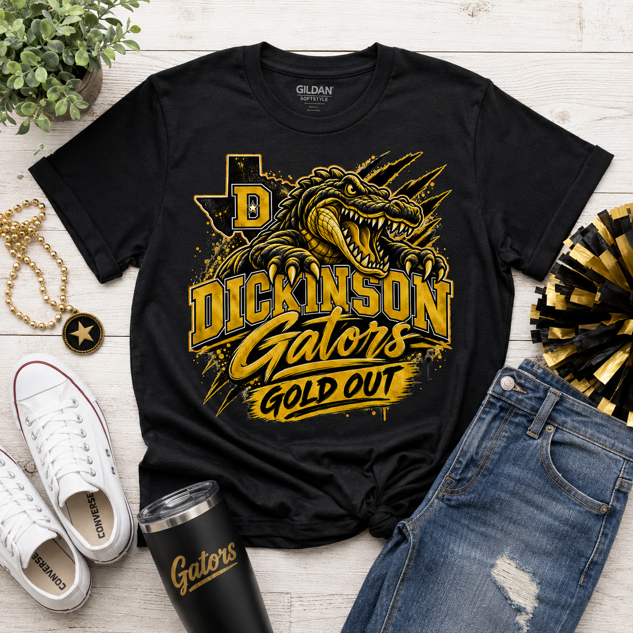 A black Gildan t-shirt with a gold and yellow graphic of a gator and the text 'DICKINSON Gators GOLD OUT', styled in a flat lay with white sneakers, blue jeans, a black tumbler, a gold chain, and a plant on a white wood background.