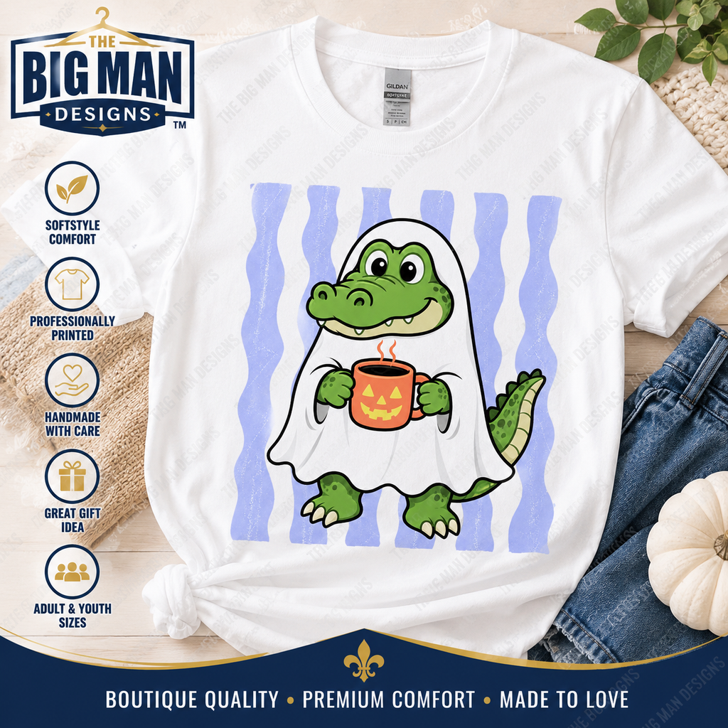 A white graphic t-shirt featuring a cartoon green alligator dressed as a ghost holding an orange pumpkin mug, set against a background of light blue and white stripes. The shirt is styled in a flat-lay arrangement with denim jeans, a small pumpkin, and a burlap sack, with branding and product feature icons from 'The Big Man Designs' visible.