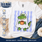 A white graphic t-shirt featuring a cartoon green alligator dressed as a ghost holding an orange pumpkin mug, set against a background of light blue and white stripes. The shirt is styled in a flat-lay arrangement with denim jeans, a small pumpkin, and a burlap sack, with branding and product feature icons from 'The Big Man Designs' visible.