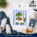 A white graphic T-shirt laid flat on a light surface with jeans, white sneakers, a coffee cup, a small book, pumpkins, and a plant around it. The shirt features a cartoon green alligator wearing a white ghost and holding a jack-o'-lantern mug, with vertical light blue stripes behind it.