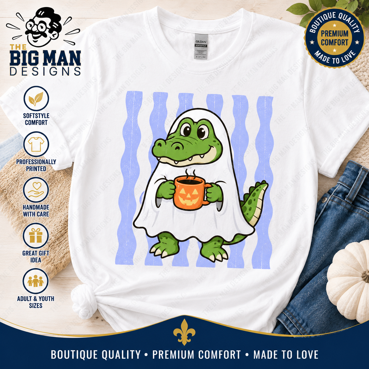 A white short-sleeve graphic T-shirt laid flat with a cartoon green alligator wearing a white ghost and holding an orange pumpkin mug, set against blue wavy stripes. The shirt is styled with denim, a small white pumpkin, and promotional badges from The Big Man Designs.