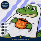 A close-up of a printed fabric design featuring a green alligator wearing a white ghost and holding an orange pumpkin mug. The image includes promotional text for a DTF print, highlighting vibrant colors and crisp detail.