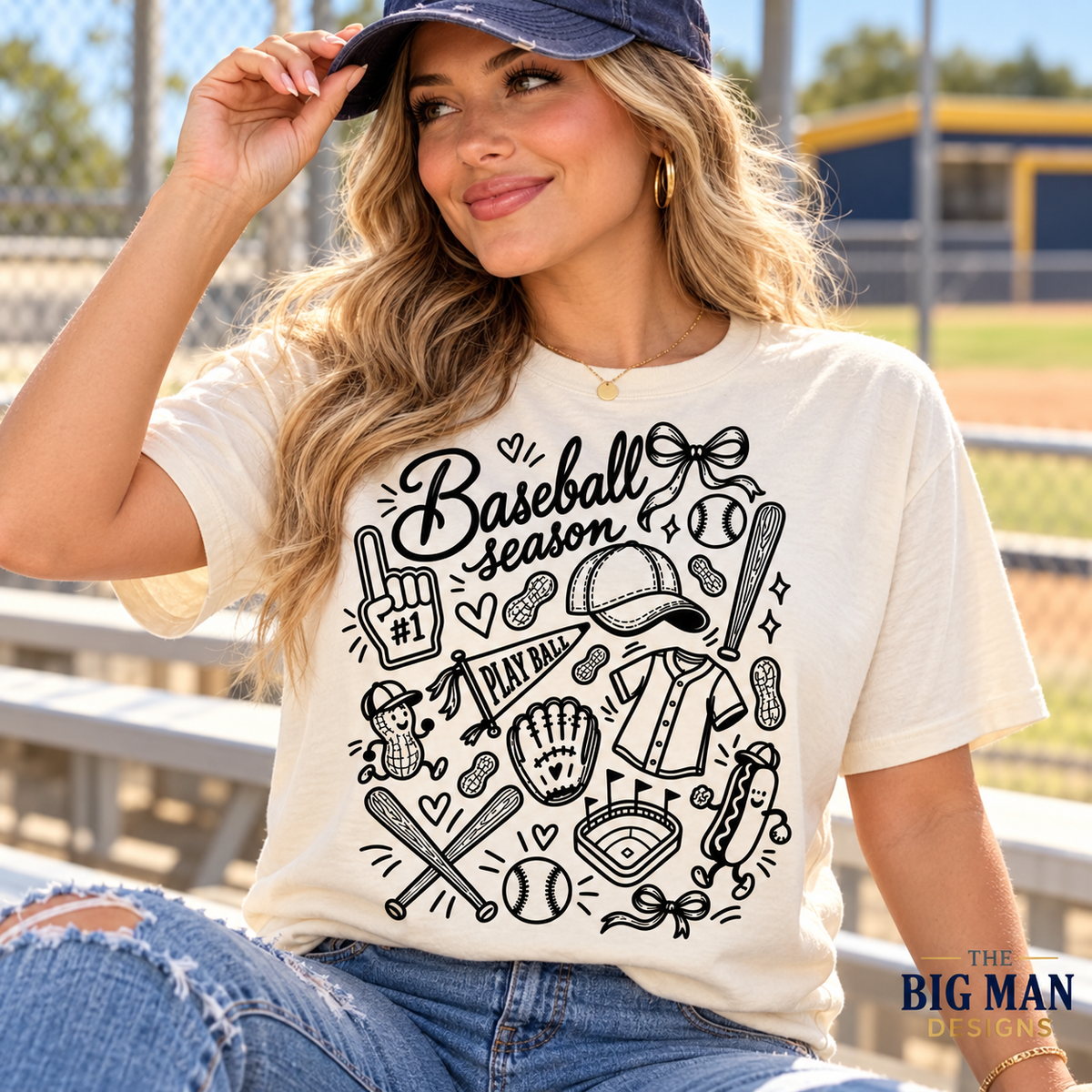 Woman wearing a natural-colored Baseball Season T-shirt with black baseball doodles and bows at a ballpark.