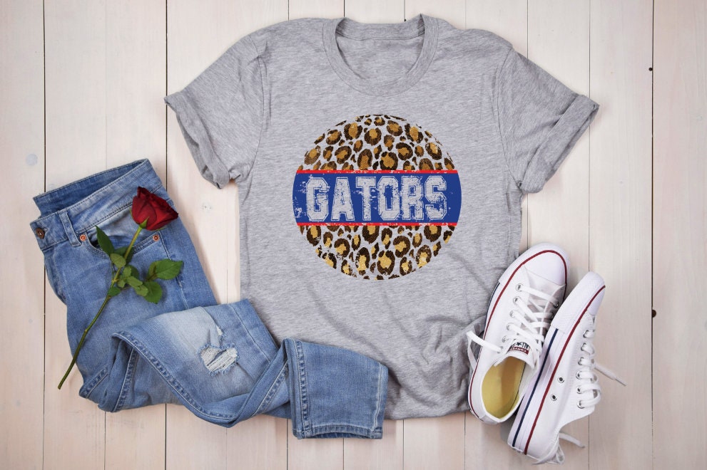 A gray t-shirt with a distressed, circular design featuring a leopard print pattern and the word 'GATORS' in blue text with a red stripe above it. The t-shirt is displayed on a white wooden surface with blue jeans and white sneakers.