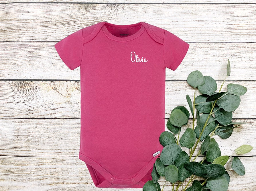 A pink baby onesie with short sleeves and a white embroidered name 'Olivia' on the front.
