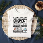 May include: A tan sweatshirt with black text that reads 'SUSPECT CLAIMS SHE LIKES WORKING WITH STUDENTS BUT IS ALWAYS WRITING REPORTS' with a checkered pattern above and below the text. The text is surrounded by a black border.