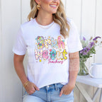 Woman wearing a white t-shirt featuring colorful “Bruh We Out Teachers” graphic, standing in front of a white background with flowers.
