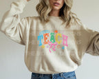 Woman wearing a natural-color sweatshirt featuring colorful "TEACH" graphic with bow and stars.