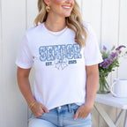 White T-shirt featuring "Senior Est. 2025" in floral blue print with a bow design, styled with jeans.
