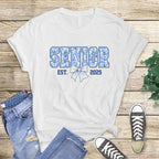 Flat lay of white Senior Est. 2025 shirt styled on wicker basket with pine accents, cozy holiday vibe.