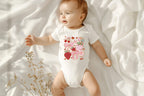 Baby laying on white sheets wearing strawberry summer onesie, soft lighting with dried flowers.