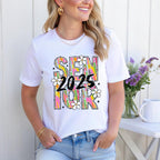 Woman smiling in white Brushstroke Senior 2025 shirt, styled with gold jewelry and jeans outdoors.
