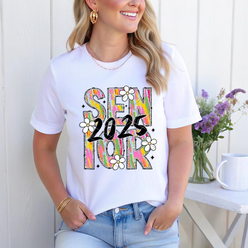 Woman smiling in white Brushstroke Senior 2025 shirt, styled with gold jewelry and jeans outdoors.