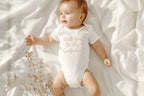 Baby lying on soft white blanket wearing 'Will You Be My Godfather?' onesie, soft natural light.