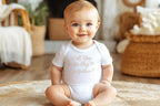 Smiling baby sitting up wearing 'Will You Be My Godfather?' onesie in cozy boho-style room