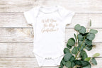 Flat lay of baby onesie reading 'Will You Be My Godfather?' with eucalyptus branch, light wood background.