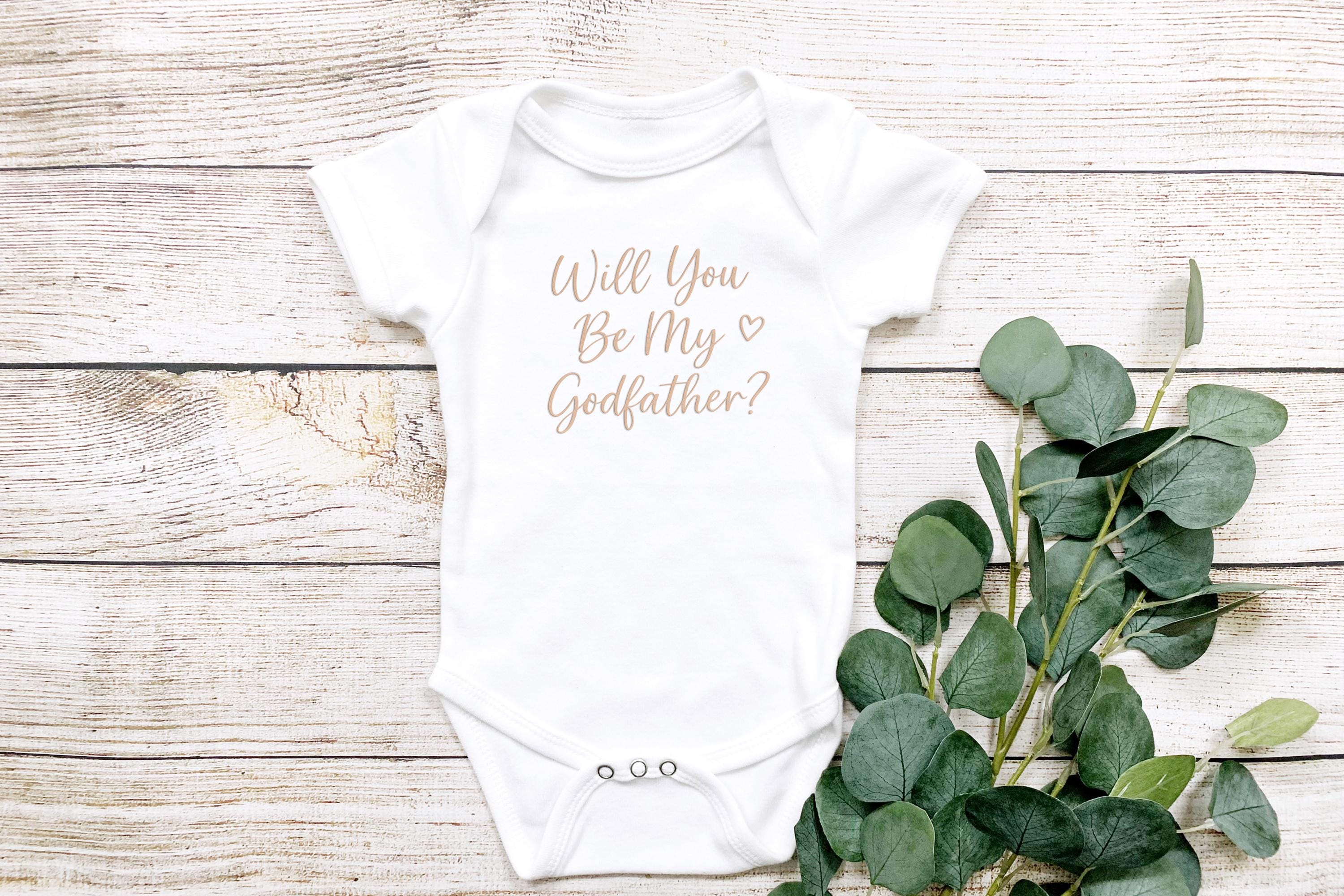 Flat lay of baby onesie reading 'Will You Be My Godfather?' with eucalyptus branch, light wood background.