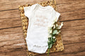 Flat lay of a white baby onesie with 'Will You Be My Godfather?' text, styled with flowers on rustic wood.