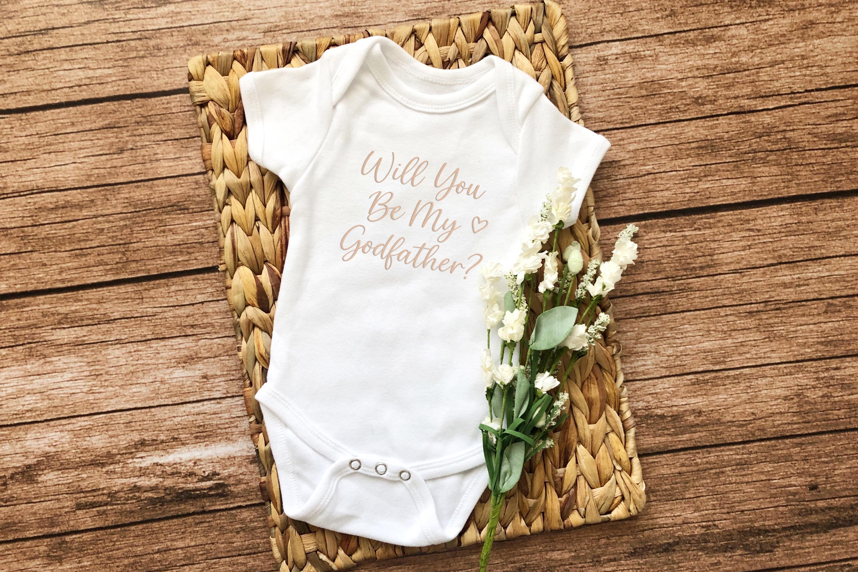 Flat lay of a white baby onesie with 'Will You Be My Godfather?' text, styled with flowers on rustic wood.