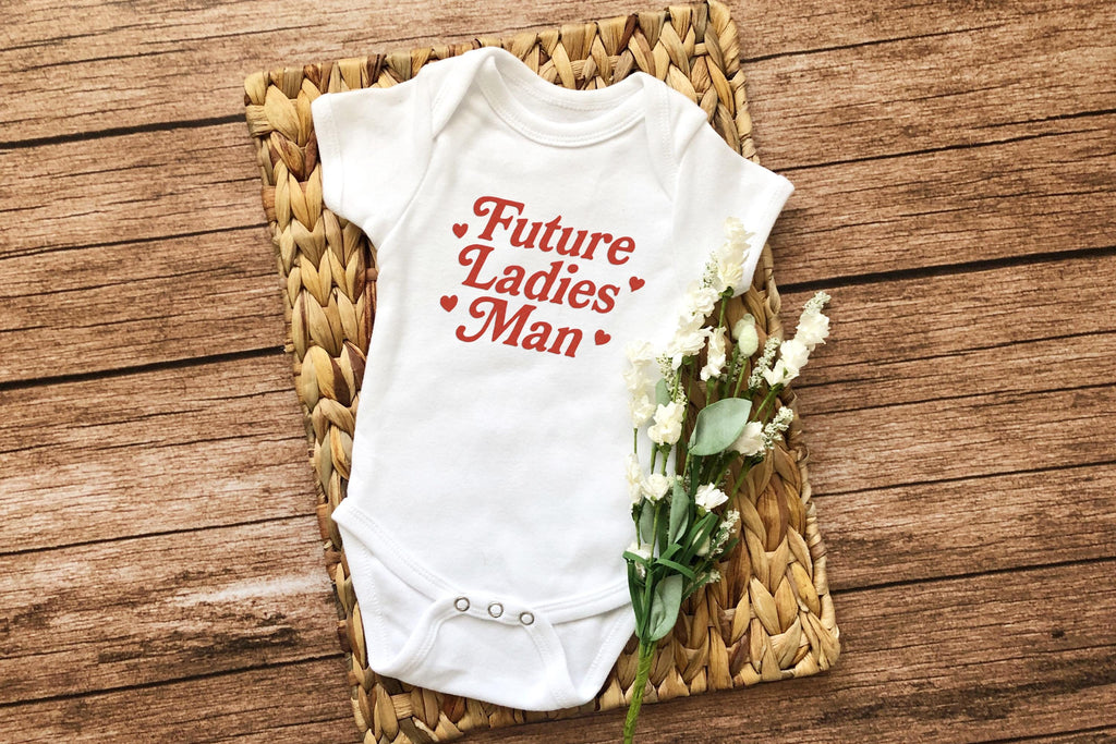 Flat lay of "Future Ladies' Man" onesie with woven basket and flowers