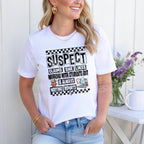 "Woman smiling and wearing a white T-shirt with a funny education-related design that says 'Suspect Claims She Likes Working With Students But Is Always Writing Reports,' standing outdoors next to purple flowers."