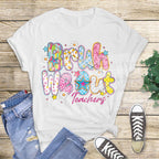 Flat lay image of a white t-shirt with colorful “Bruh We Out Teachers” print, styled with jeans and sneakers on a rustic wood background.
