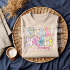 Folded natural-color t-shirt featuring “Bruh We Out Teachers” vibrant design, placed in a wicker basket with a cozy background.
