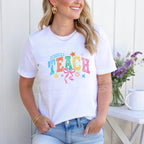 Woman standing wearing a white T-shirt with colorful "TEACH" design, smiling.