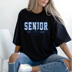 Black oversized T-shirt with "Senior Est. 2025" blue floral lettering and bow, worn casually with ripped jeans.