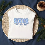 Flat lay of white Senior Est. 2025 shirt styled on wicker basket with pine accents, cozy holiday vibe.