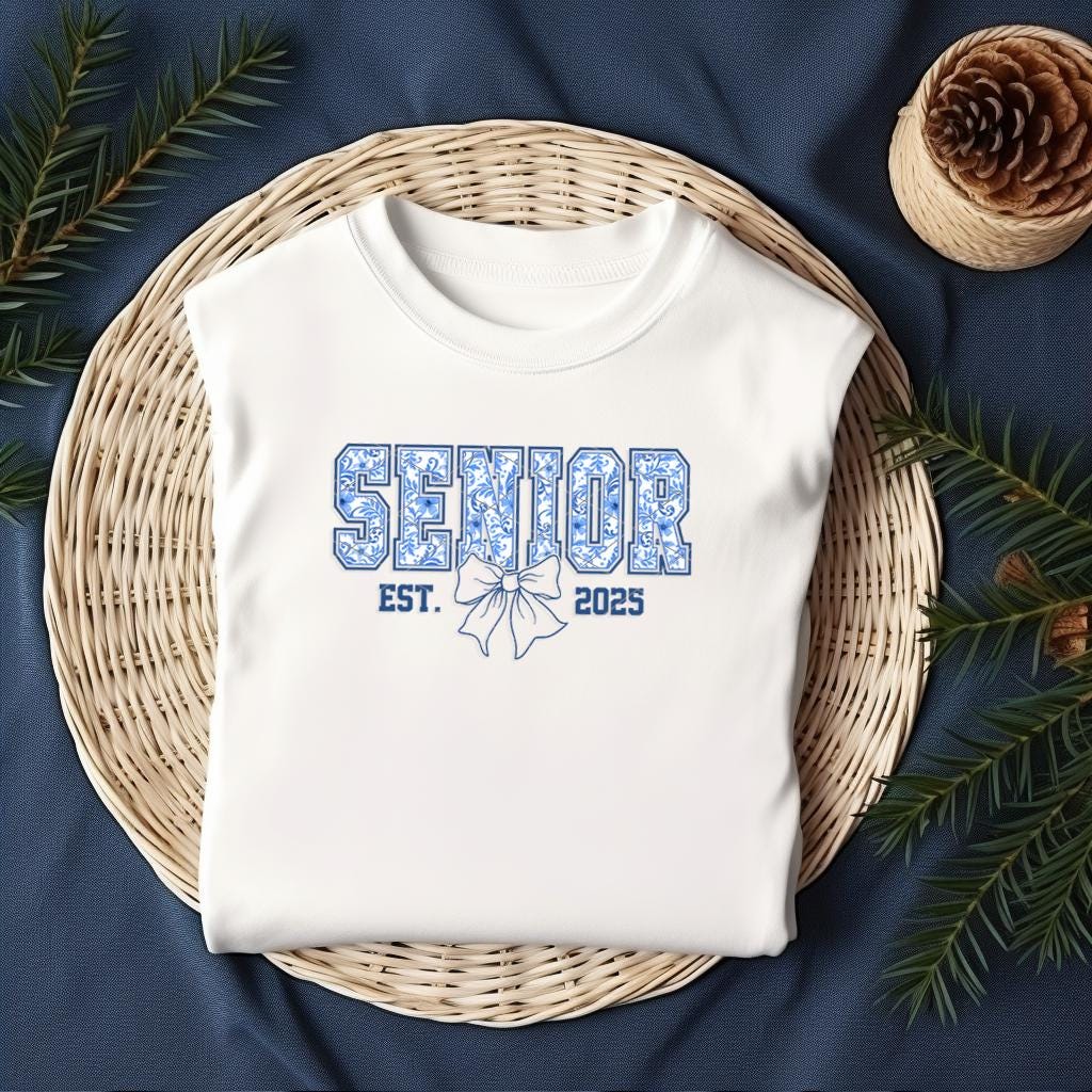 Flat lay of white Senior Est. 2025 shirt styled on wicker basket with pine accents, cozy holiday vibe.