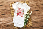 Baby onesie with strawberry and flower design, laid on woven mat with white flowers.
