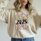 Woman wearing sand-colored sweatshirt with colorful Brushstroke Senior 2025 design and casual jeans.