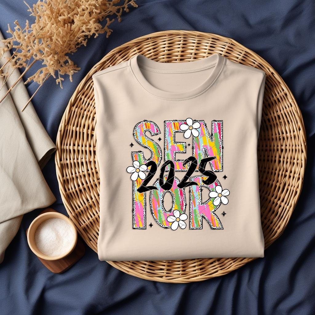 Flat lay of natural color Brushstroke Senior 2025 shirt in a wicker basket on navy background with cozy