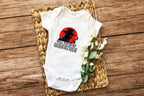 Flat lay of white baby onesie with mafia-style silhouette and godfather text, styled with wicker tray and flowers.
