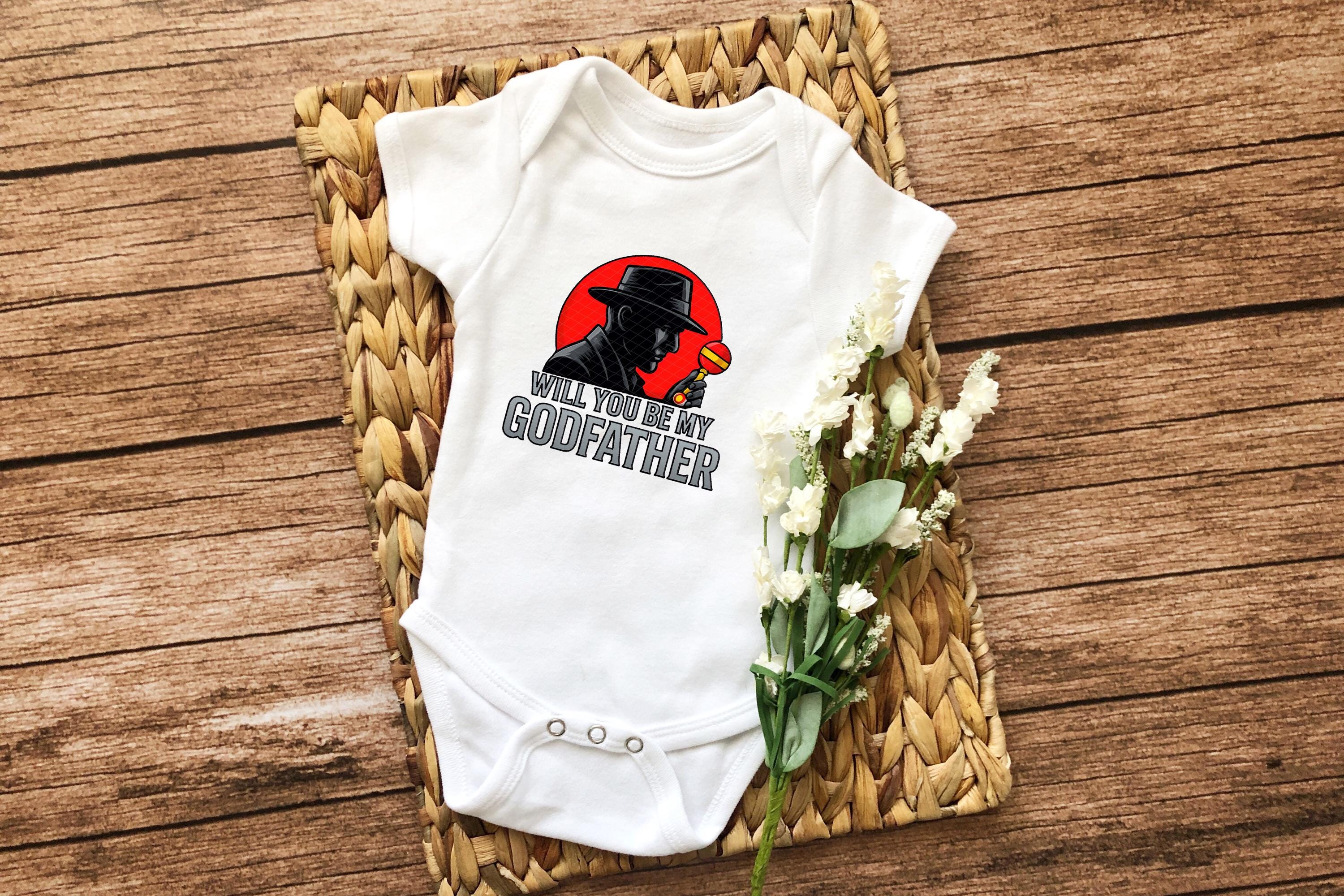 Flat lay of white baby onesie with mafia-style silhouette and godfather text, styled with wicker tray and flowers.