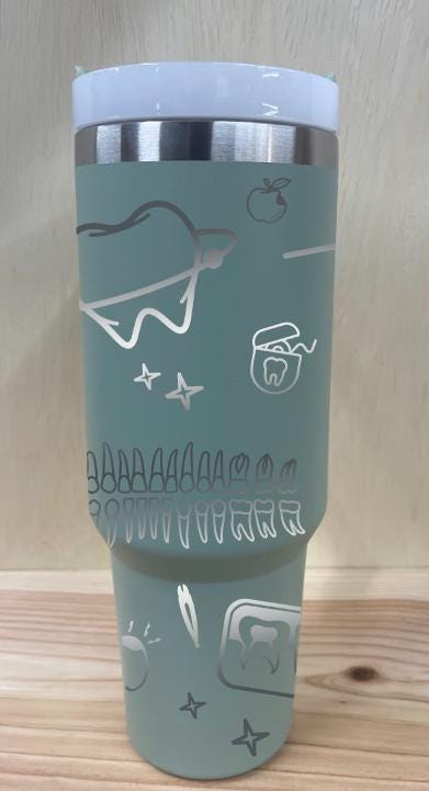 May include: A sage green stainless steel travel mug with a sage green handle and a clear lid. The mug has a silver design engraved into it.
