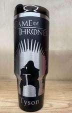 May include: Black stainless steel travel mug featuring Game of Thrones. The mug has a silver design of a dragon, a wolf, and the words "House Lannister", "Mother of Dragons", and "House Baratheon".