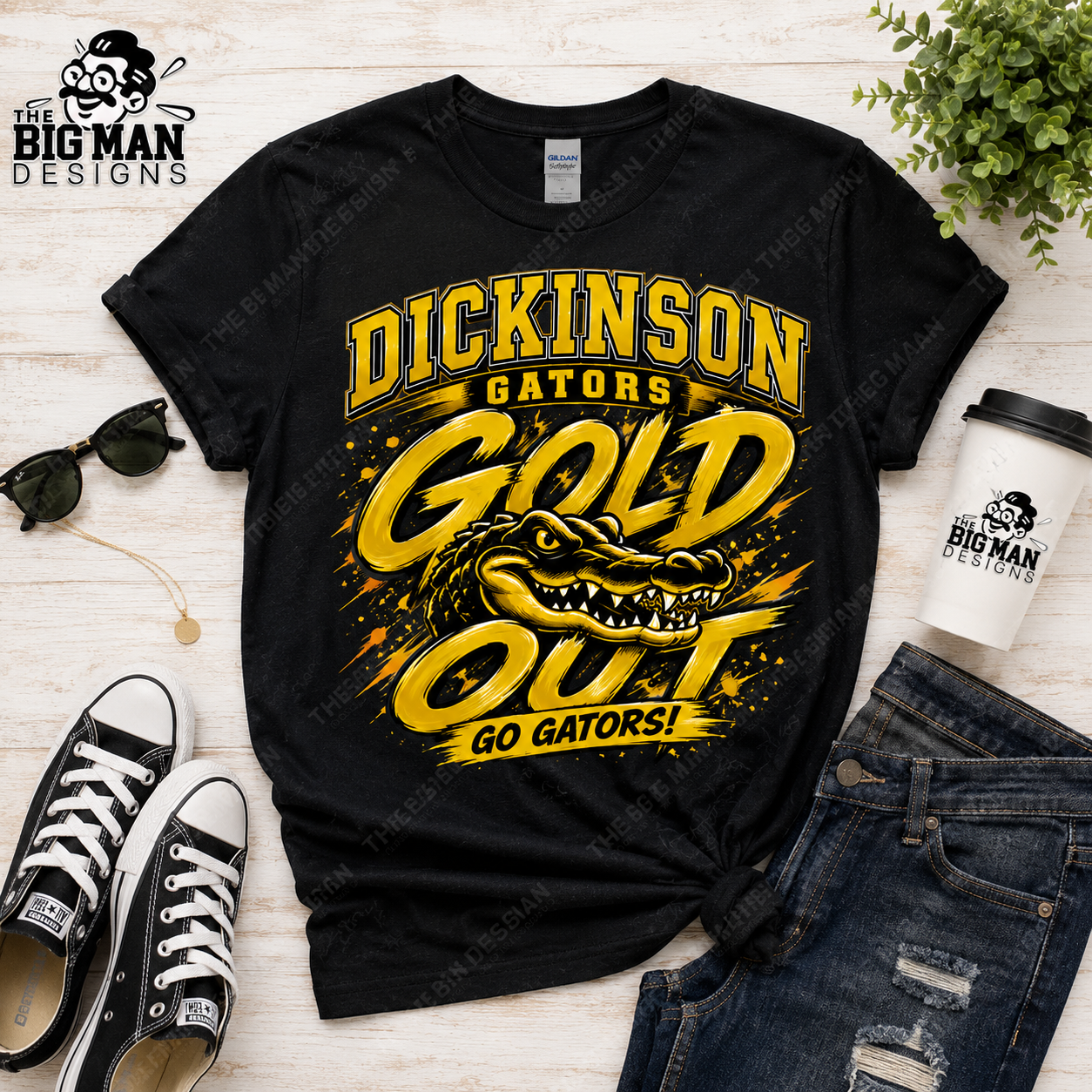 A black graphic t-shirt featuring a yellow and gold 'Dickinson Gators Gold Out Go Gators!' design with an alligator illustration, styled in a flat lay with jeans, sneakers, sunglasses, a coffee cup, and a plant on a light wood background.
